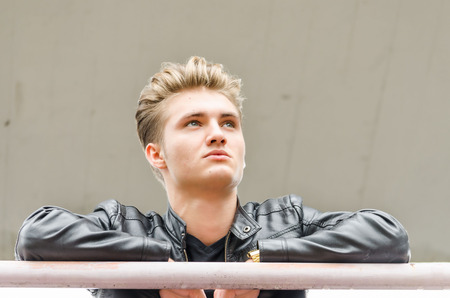 Attractive young man wearing black leather jacket looking outside from balconyの写真素材