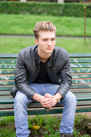 Handsome blond young man sitting on green, wooden park benchの写真素材