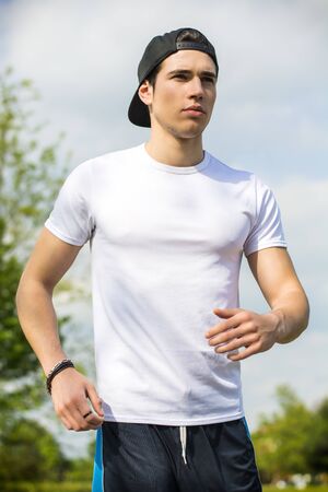 Handsome young man running and jogging on road in the country in a sunny day, wearing white shirt and baseball capの写真素材