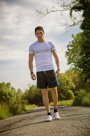 Handsome young man walking and trekking on road in the country in a sunny day, wearing white shirt and baseball capの写真素材