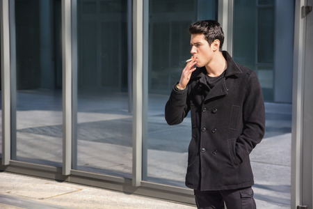 Handsome stylish young man smoking outside in urban setting, looking awayの写真素材
