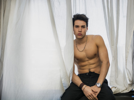 Handsome fit athletic young man shirtless, sitting on window ledge looking at cameraの写真素材