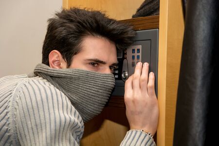 Young male burglar hiding face with mask opening a small home vault or safe