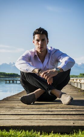 Handsome young man on a lake in a sunny, peaceful day, sitting on a wood pier, thinking or meditatingの写真素材