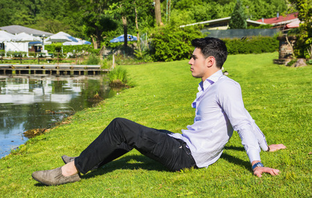 Handsome young man sitting on lawn nexto to a lake in a sunny, peaceful day, looking away to a sideの写真素材