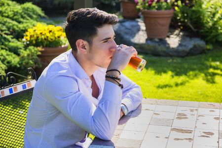 Handsome young man sitting alone at table outside in park or nature, with glass of soda beverageの写真素材