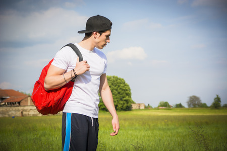Handsome young man outdoor hiking on rural road with backpack on shoulder, looking at country around himの写真素材