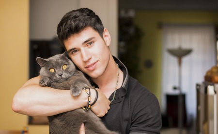 Handsome Young Animal-Lover Man Inside the House, Hugging his Gray Domestic Cat Pet.の写真素材