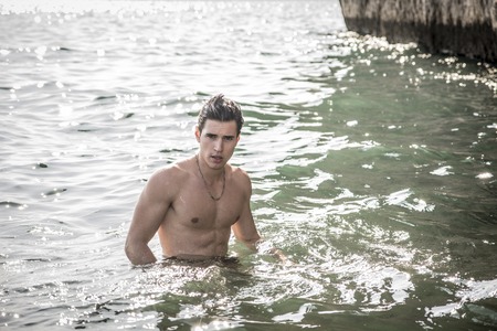 Attractive young shirtless athletic muscle man standing in water by sea or ocean shore, wearing shorts, showing muscular bodyの写真素材