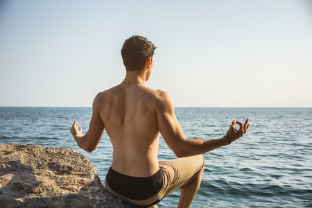 Handsome Shirtless Young Man During Meditation or Doing an Outdoor Yoga Exercise Sitting on Rocks by...
