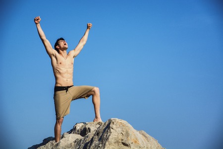 Successful sexy muscular shirtless young man with fists raised, shouting for joy, happy, standing on big rock with blue sky behind himの写真素材