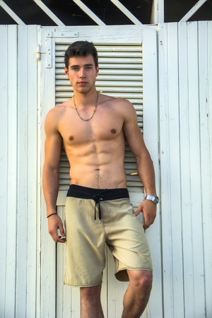 Handsome and muscular athletic shirtless young man standing against white beach changing room wall, looking confident at cameraの写真素材