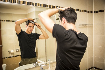 Reflection of Young Man Bushing Hair in Bathroom Mirror Getting Ready to Go Outの写真素材