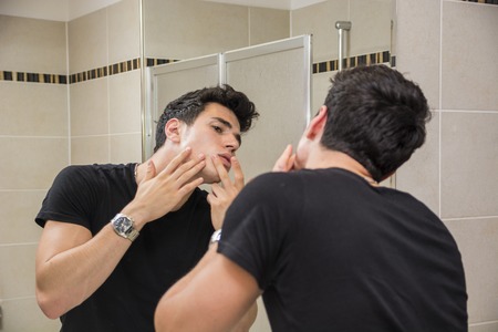 Handsome Young Man in Bathroom Touching his Face, Squeezing a Spot or Examining Blemishesの写真素材