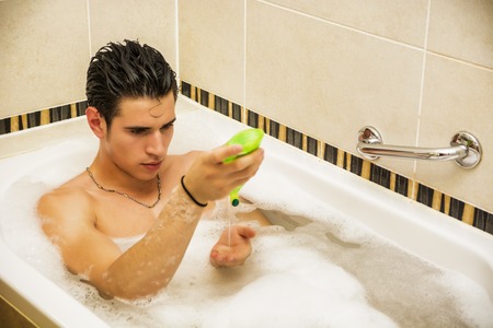 Handsome young man in bathtub at home having bath, washing body and hair with bathfoam and shampooの写真素材