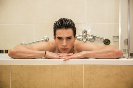 Handsome young man in bathtub at home having bath, leaning on tub edge, looking at cameraの写真素材
