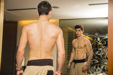 Young Shirtless Handsome Muscular Man Admiring His Muscles in Gym Mirror or Checking Progress and Training Results, as seen from Behindの写真素材