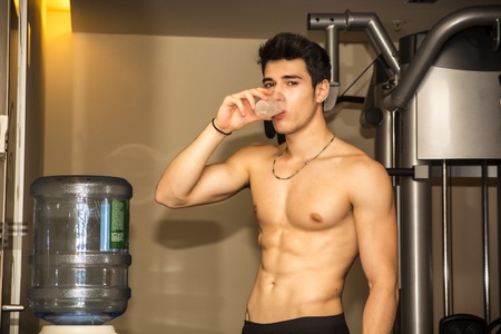 Attractive athletic shirtless young man drinking water from dispenser in gym while looking at cameraの写真素材