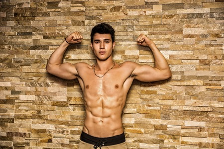 Attractive and muscular shirtless young man leaning against stone wall doing double bicep poseの写真素材