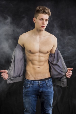 Attractive trendy athletic fit young man undressing, taking off t-shirt, on dark smoky backgroundの写真素材