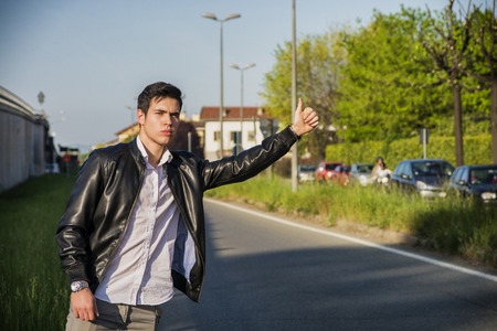 Handsome young man, a hitchhiker waiting for car on roadside in city, wearing black leather jacketの写真素材