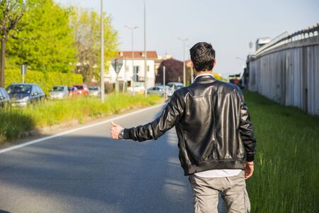 Back of young man, a hitchhiker waiting for car on roadside in city, wearing black leather jacketの写真素材