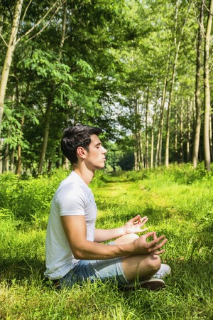 Handsome Young Man During Meditation or Doing an Outdoor Yoga Exercise Sitting Cross Legged on Grassy Ground Alone in Woodsの写真素材