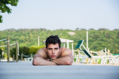 Attractive Young Dark Haired Man Lying on Stomach on Sun Deck with Lounge Chairs, Outdoors on Sunny Day with Forest in Backgroundの写真素材