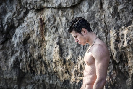 Muscular young shirtless athletic man standing in water by sea or ocean shore,  looking down, with rocks behind himの写真素材