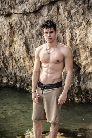 Attractive muscular young shirtless athletic man standing in water by sea or ocean shore, wearing shorts, looking at camera, with rocks behind himの写真素材