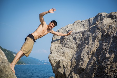 Handsome Shirtless Young Man Wearing Swim Shorts and Waving at Camera Leaning Across Gap Between Two Massive Boulders at Picturesque Ocean Coastの写真素材