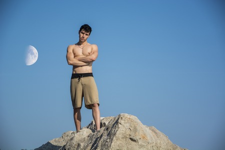 Muscular young man shirtless standing on rock against the sky with moon next to him, seen from below perspectiveの写真素材