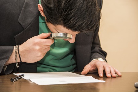 Young Man Sitting at Desk Scrutinizing Paper Contract with Magnifying Glass Prior to Signing