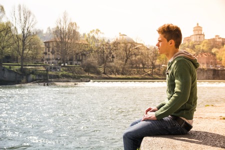 Three-quarter length of contemplative light brown haired teenage boy wearing green hooded-shirt and denim jeans sitting on wall beside picturesque river in Turin, Italyの写真素材