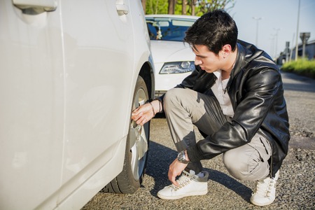 Handsome Young Man in Casual Clothing Squatting Besides the Wheel of a his White Car Changing Tiresの写真素材