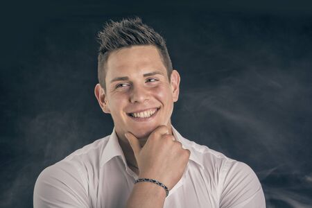 Headshot of smiling attractive young man in white shirt on dark backgroundの写真素材
