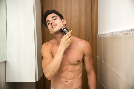 Muscular man shirtless using electric shaver, looking at camera smiling, in his bathroomの写真素材