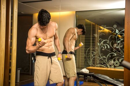 Funny young muscular shirtless man working out in gym with ridiculous, tiny dumbbells, lifting...