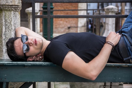 Attractive young man laying down on wood bench in European city, wearing sunglasses