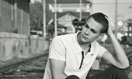 Attractive young man sitting on railroad, wearing polo shirt, looking away, black and white shotの写真素材