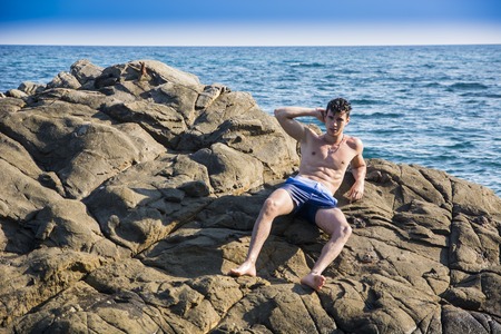Attractive young shirtless athletic man laying down on rock by water on ocean or sea shore, wearing...