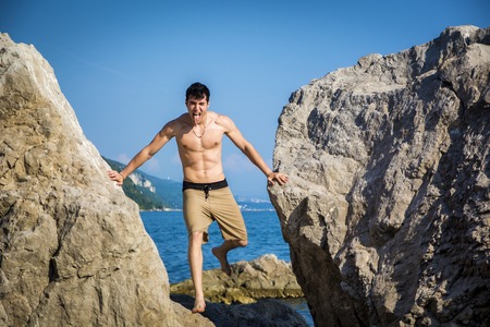 Handsome Shirtless Young Man Wearing Swim Shorts Leaning Across Gap Between Two Massive Boulders at Picturesque Ocean or Sea Coastの写真素材