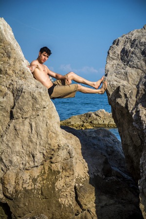 Handsome Shirtless Young Man Wearing Swim Shorts Leaning Across Gap Between Two Massive Boulders at Picturesque Ocean or Sea Coastの写真素材