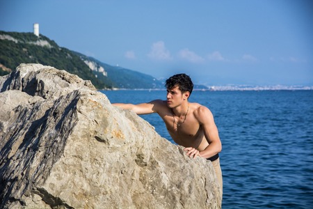 Attractive young shirtless athletic man climbing on rock by water on ocean or sea shore, wearing shorts, looking at cameraの写真素材