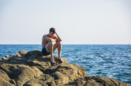 Muscular young man shirtless sitting on rock with sky and sea or ocean behind. Large copyspaceの写真素材
