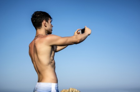 Side View of a Shirtless Young Man Taking Selfie Photos at the Beach While Sitting on Big Rock Under the Heat of the Sun.の写真素材