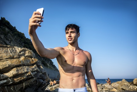 Side View of a Shirtless Young Man Taking Selfie Photos at the Beach While Sitting on Big Rock Under the Heat of the Sun.の写真素材