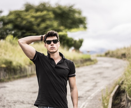 Sexy handsome young man in jeans and black t-shirt walking along rural road, with sunglassesの写真素材