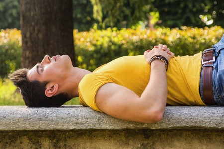 Attractive young man sleeping on stone bench outdoor in city park during dayの写真素材