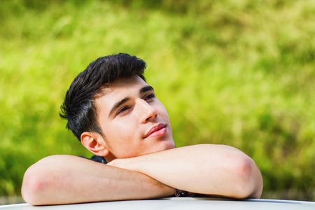 Head and arms shot of handsome attractive young man looking at camera outdoor, leaning on flat surface with head resting on handsの写真素材